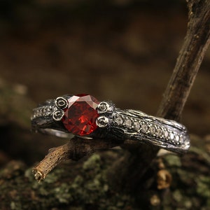 May include: A silver ring with a dark red gemstone and small white gemstones set in a textured band. The ring is resting on a branch.