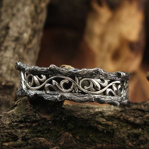 May include: A silver wedding band featuring an intricate design of interwoven branches and leaves. The band is displayed against a rustic wooden backdrop.