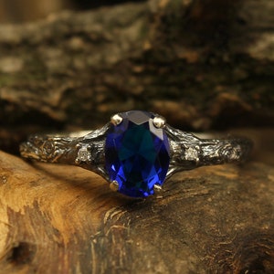 May include: A silver ring with a dark blue oval gemstone and smaller diamonds on the band. The ring is set on a piece of wood.