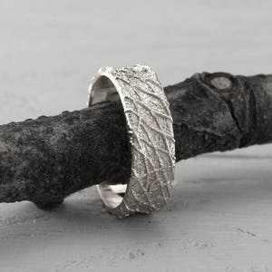May include: A silver wedding band with a textured, organic pattern. The band is resting on a piece of driftwood.