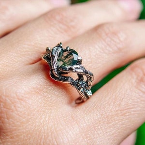 May include: A silver ring with a green gemstone set in a branch-like design. The ring is on a hand with a green background.