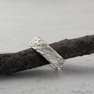 May include: A silver wedding band with a textured, organic design. The band is shown resting on a piece of driftwood.