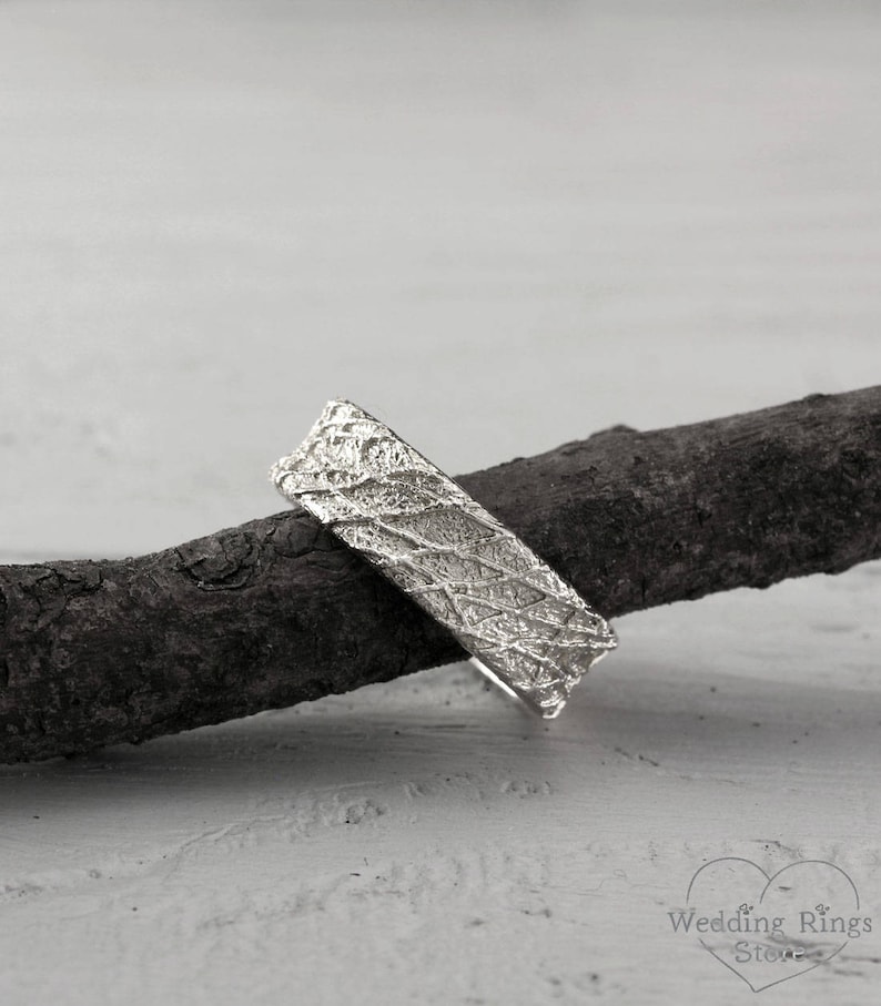 May include: A silver wedding band with a textured, organic design. The band is shown resting on a piece of driftwood.