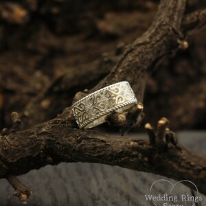 May include: A silver wedding band with a delicate, intricate design featuring a repeating pattern of hearts and swirls. The band is wide and has a slightly rounded edge.