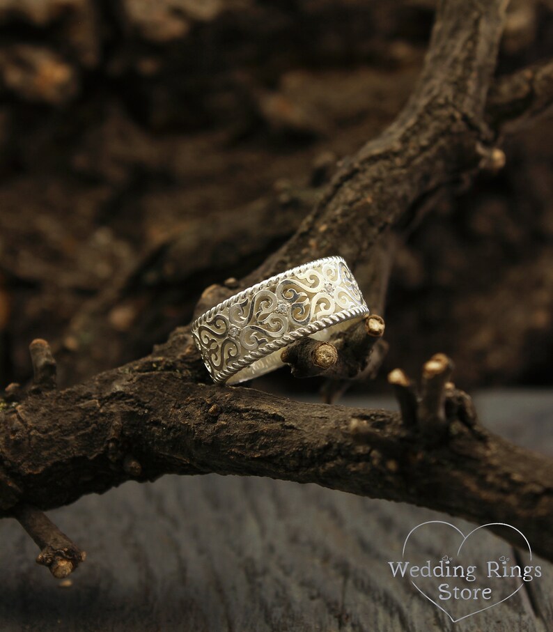 May include: A silver wedding band with a delicate, intricate design featuring a repeating pattern of hearts and swirls. The band is wide and has a slightly rounded edge.