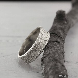 May include: A silver wedding band with a textured, organic pattern. The band is resting on a piece of driftwood.