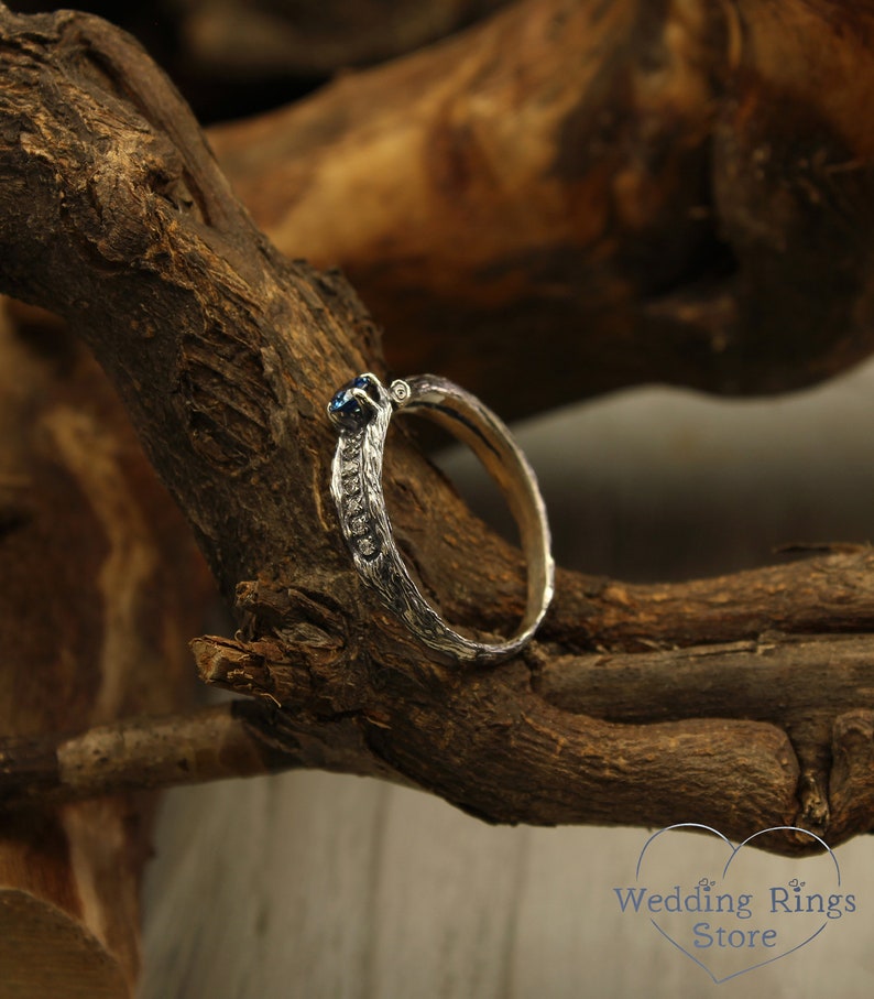 May include: A silver wedding band with a blue gemstone set in the center. The band has a textured, organic design.