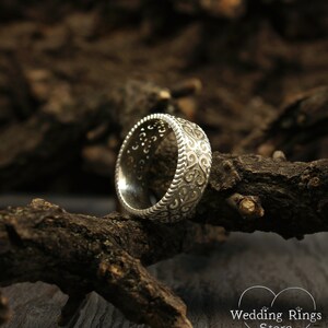 May include: A silver wedding band with a detailed, intricate design. The band is on a piece of wood.