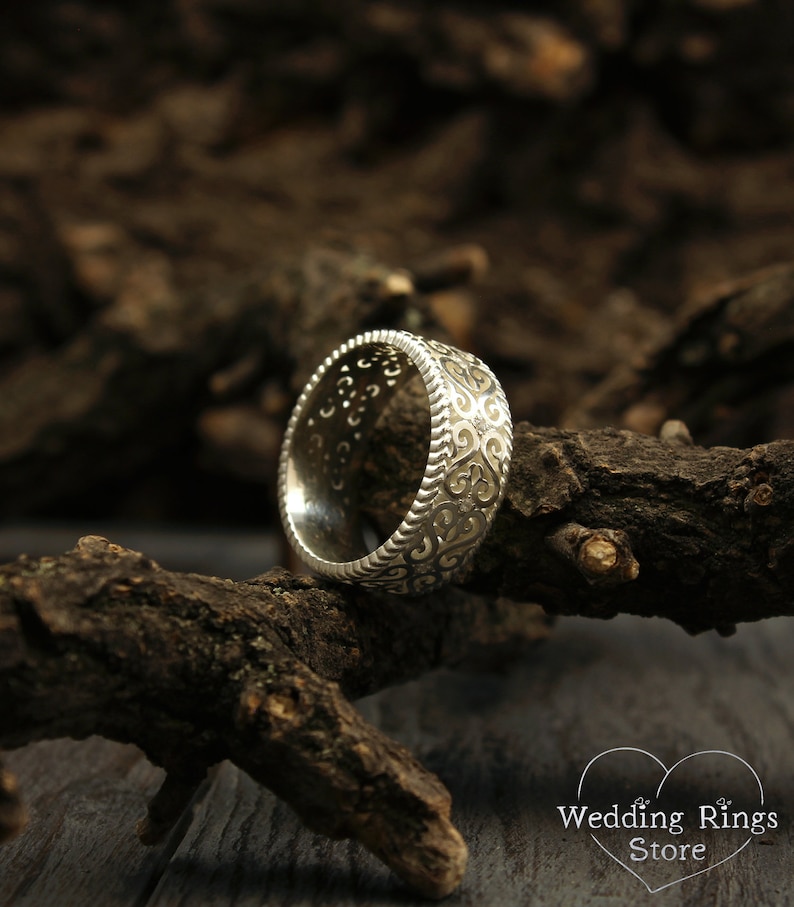 May include: A silver wedding band with a detailed, intricate design. The band is on a piece of wood.