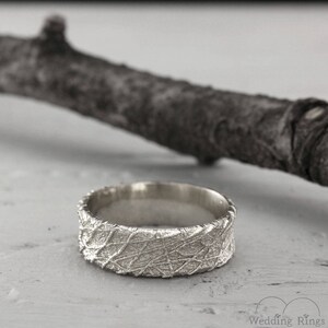 May include: A silver wedding band with a textured, organic pattern. The band is wide and has a rounded profile.