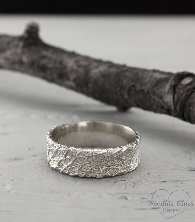 May include: A silver wedding band with a textured, organic pattern. The band is wide and has a rounded profile.