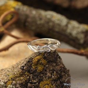 May include: A silver wedding band with a delicate, intricate design of intertwined branches. The ring is set against a backdrop of a mossy rock.