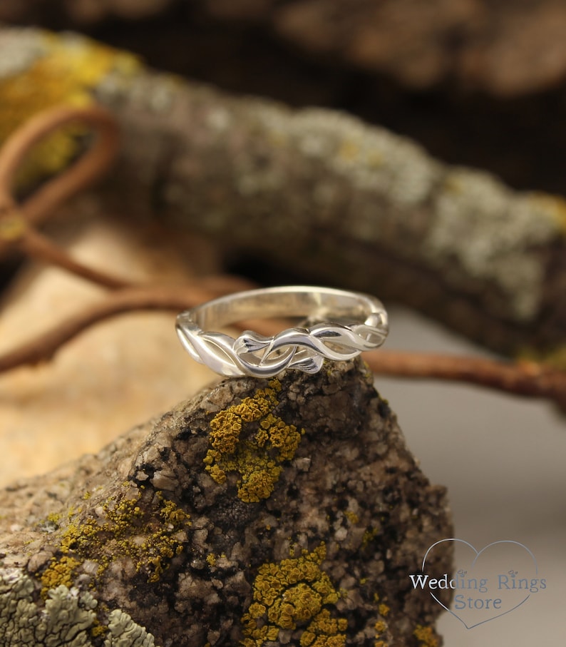 May include: A silver wedding band with a delicate, intricate design of intertwined branches. The ring is set against a backdrop of a mossy rock.