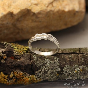 May include: A silver wedding band with a unique, intricate design featuring a twisted, interwoven pattern. The band is shown on a piece of wood with lichen.