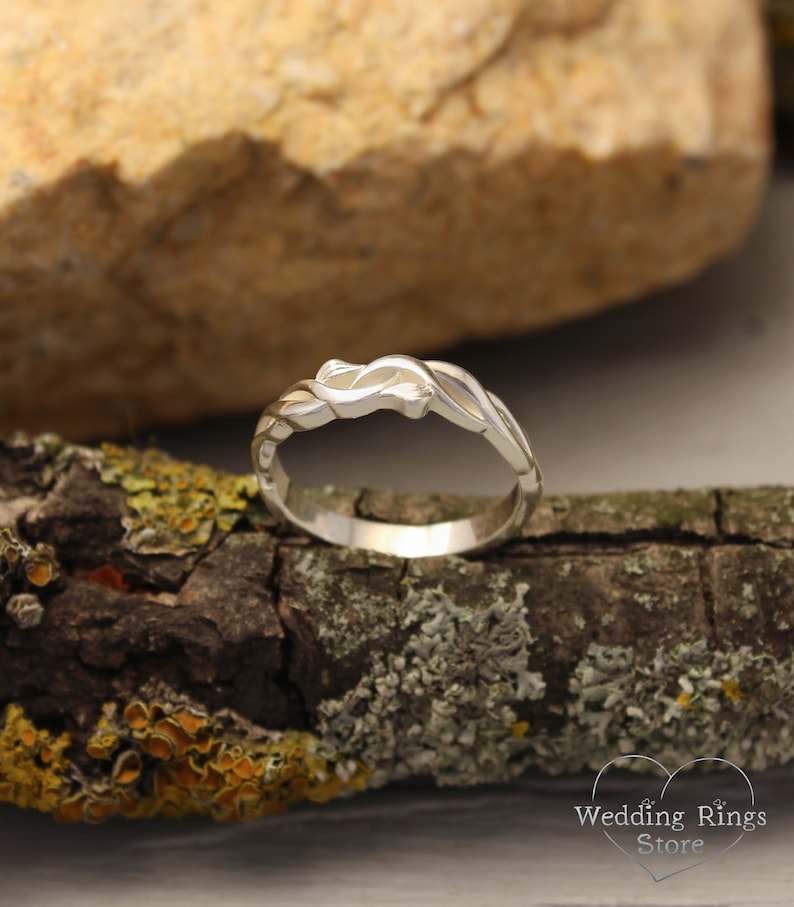 May include: A silver wedding band with a unique, intricate design featuring a twisted, interwoven pattern. The band is shown on a piece of wood with lichen.