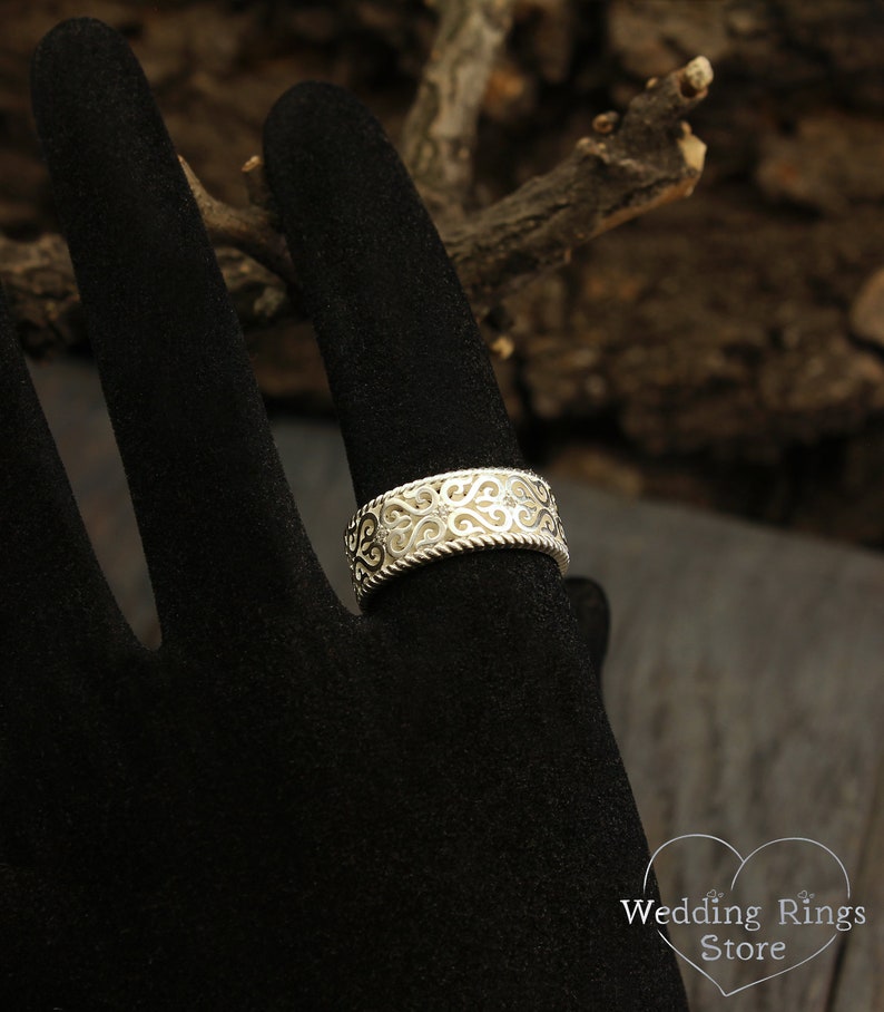 May include: A silver filigree wedding band with a twisted rope design. The band is on a black velvet ring display.