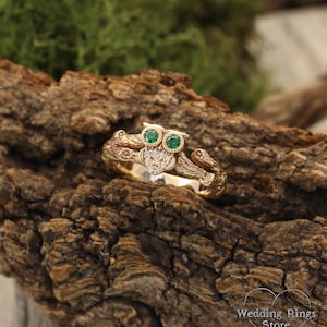 Può includere: Un anello d'oro con un designo di gufo dettagliato, occhi verde smeraldo e un corpo fatto di piccoli diamanti. L'anello è posto su un pezzo di corteccia d'albero.
