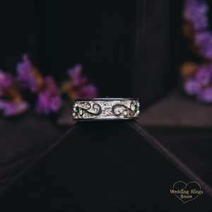 May include: A silver wedding band with a detailed, intricate design featuring a scrolling pattern and small green gemstones.