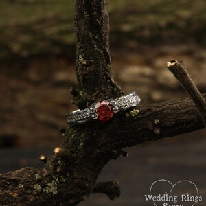 May include: A silver ring with a red gemstone set in a decorative band. The ring is resting on a branch.