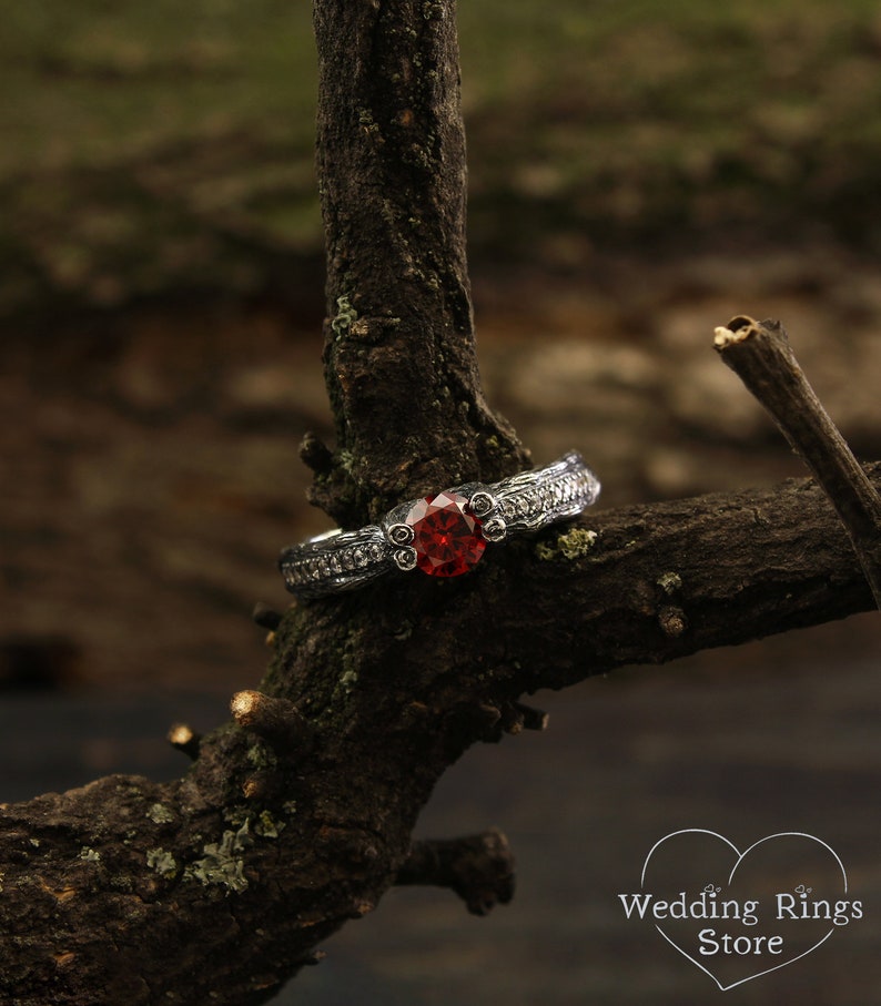 May include: A silver ring with a red gemstone set in a decorative band. The ring is resting on a branch.