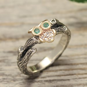 May include: A silver ring with an owl motif. The owl has green gemstone eyes and sits on a branch. The owl's body is decorated with small, clear gems, and the ring has a textured, rustic finish. The ring is a unique piece of jewellery.