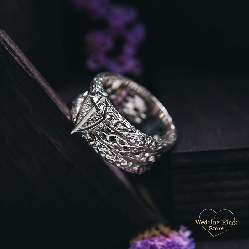 May include: A silver ring with a detailed, intricate design. The ring features a pointed, triangular shape on the top.