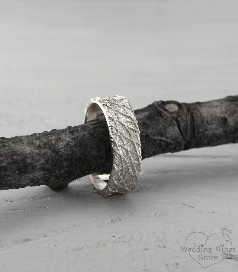 May include: A silver wedding band with a textured, organic pattern. The band is resting on a piece of driftwood.