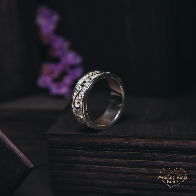 May include: A silver wedding band with a green gemstone and an intricate design. The band is on a dark wooden surface.