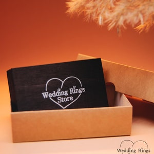 May include: A black wooden box with a white heart design and the text "Wedding Rings Store" inside. The box is open and sitting on a brown surface.