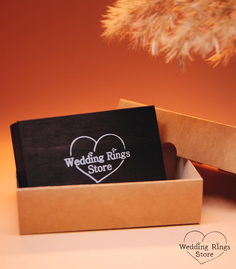 May include: A black wooden box with a white heart design and the text "Wedding Rings Store" inside. The box is open and sitting on a brown surface.