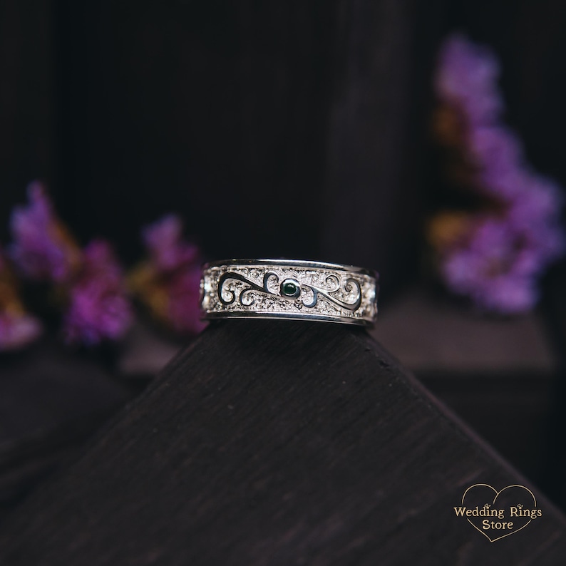 May include: A silver wedding band with a detailed scroll design and a small green gemstone.
