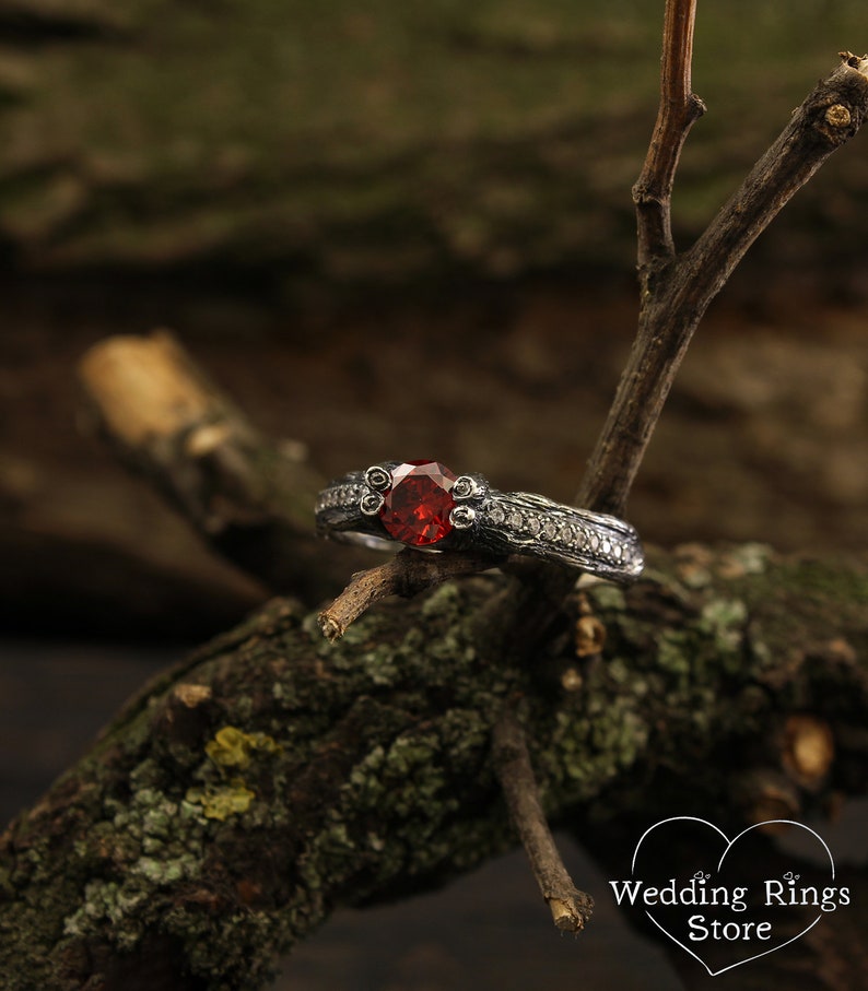 May include: A silver ring with a dark red gemstone and small white gemstones set in a textured band. The ring is resting on a branch.