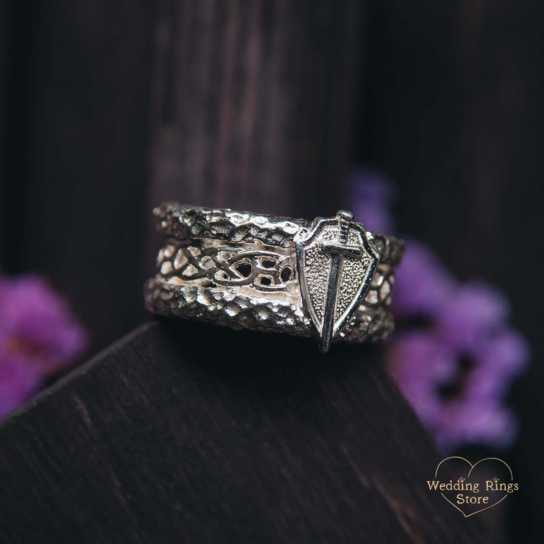 May include: A silver wedding band with a Celtic knot design and a shield with a sword on top. The band has a textured finish.