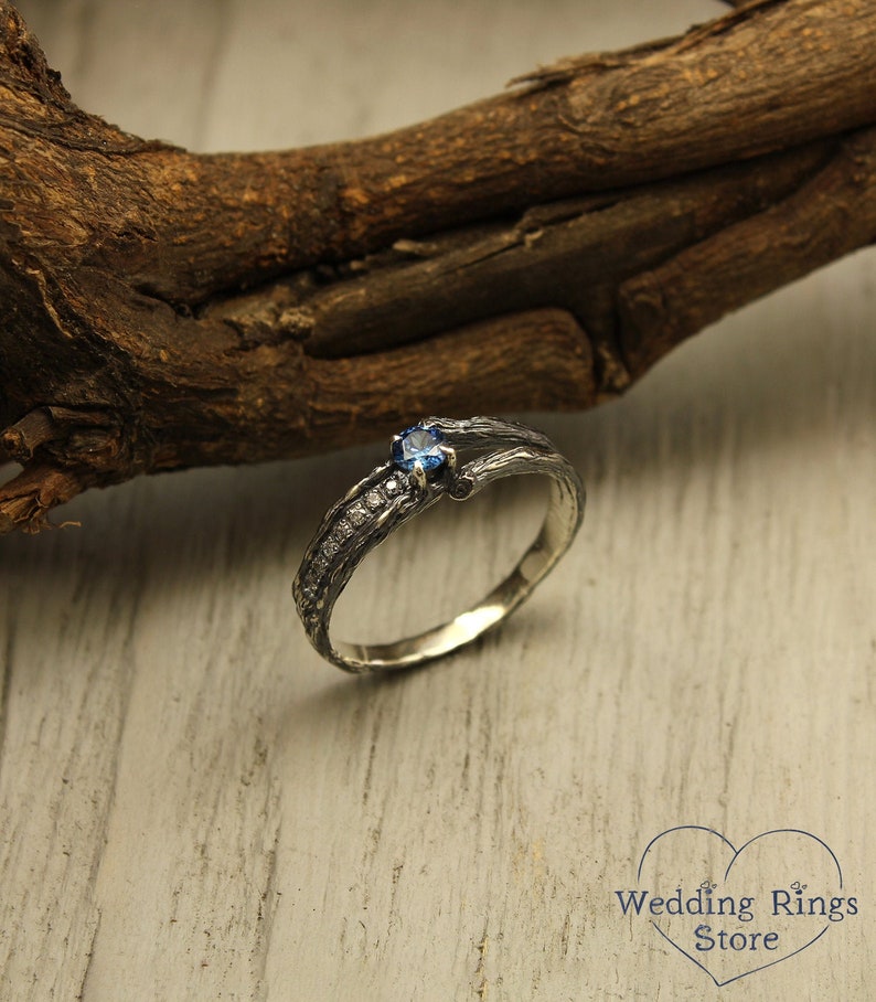 May include: A silver ring with a blue gemstone and a textured band that resembles a tree branch. The ring is set on a wooden surface.