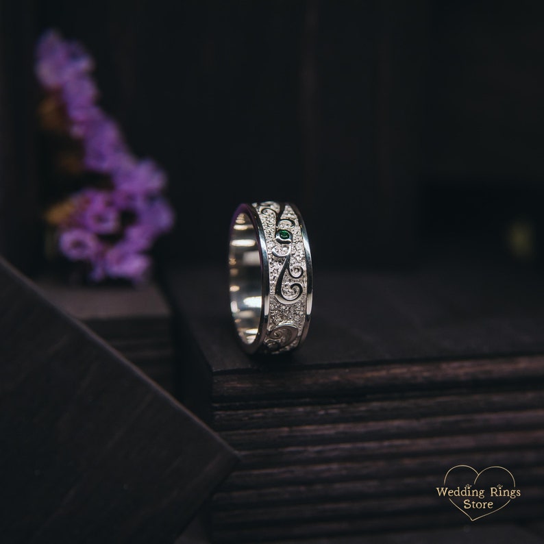 May include: A silver wedding band with a detailed design featuring a green gemstone and small white stones. The band is on a dark wooden surface.