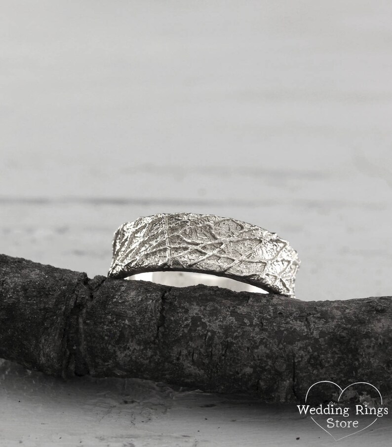 May include: A silver wedding band with a textured, organic design. The band is resting on a piece of driftwood.
