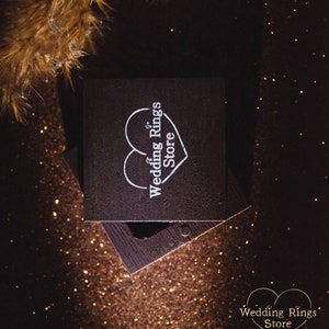 May include: A dark brown wooden ring box with a white heart and the text "Wedding Rings Store" on the lid. The box is sitting on a sparkly brown surface.