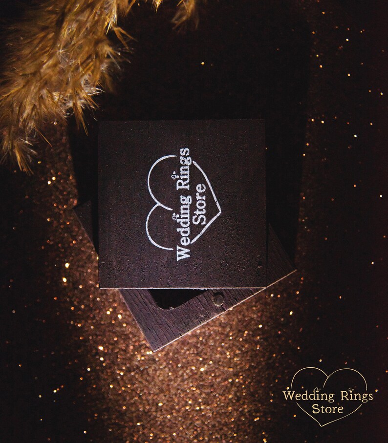 May include: A dark brown wooden ring box with a white heart and the text "Wedding Rings Store" on the lid. The box is sitting on a sparkly brown surface.