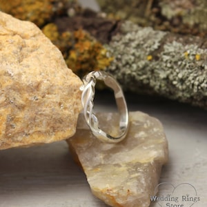 May include: A silver wedding band with a twisted, vine-like design. The band is displayed on a rock.
