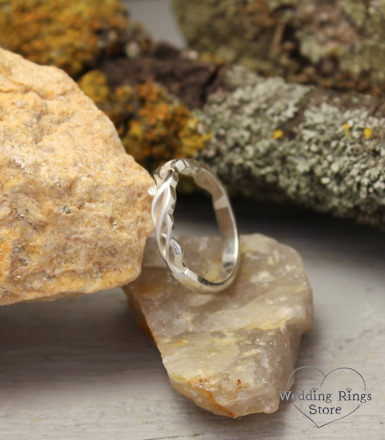 May include: A silver wedding band with a twisted, vine-like design. The band is displayed on a rock.