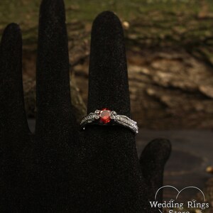 May include: A silver ring with a red gemstone set in a delicate floral design. The ring is on a black ring display stand.