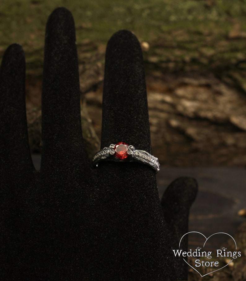 May include: A silver ring with a red gemstone set in a delicate floral design. The ring is on a black ring display stand.