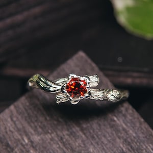 Könnte beinhalten: Ein silberner Ring mit einem roten Edelstein, der in einem Blumenmuster gefasst ist. Die Ringschiene ist so gestaltet, dass sie einem Zweig ähnelt.
