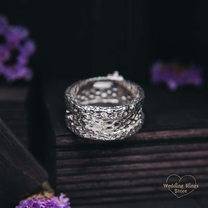 May include: A silver wedding band with a textured, organic design. The band is wide and has a unique, modern look.