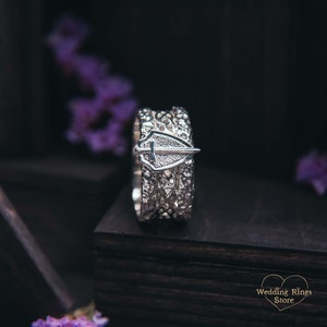 May include: A silver wedding band with a shield and sword design. The band is made of multiple layers of intricate metalwork.