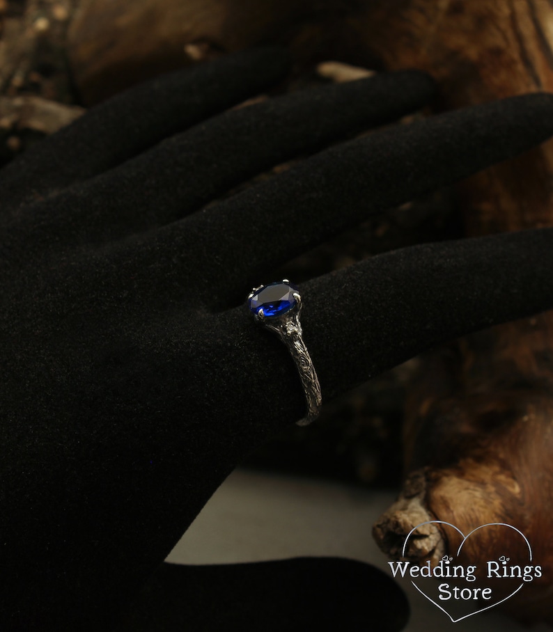 May include: A silver ring with a blue gemstone set in a claw setting. The ring band is textured and has a branch-like design.
