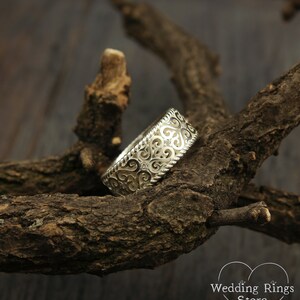 May include: A silver wedding band with a delicate, intricate design featuring swirls and hearts. The band is set against a rustic wooden background.