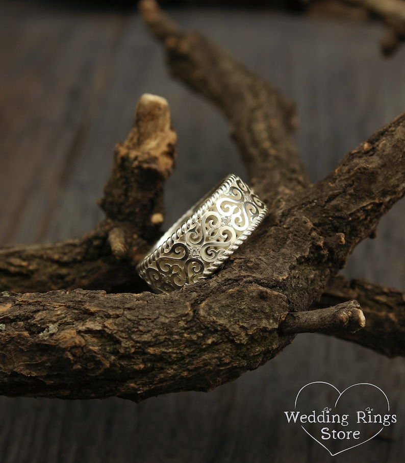 May include: A silver wedding band with a delicate, intricate design featuring swirls and hearts. The band is set against a rustic wooden background.