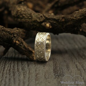 May include: A silver wedding band with a delicate, intricate design featuring a repeating heart pattern. The band is shown on a wooden surface.