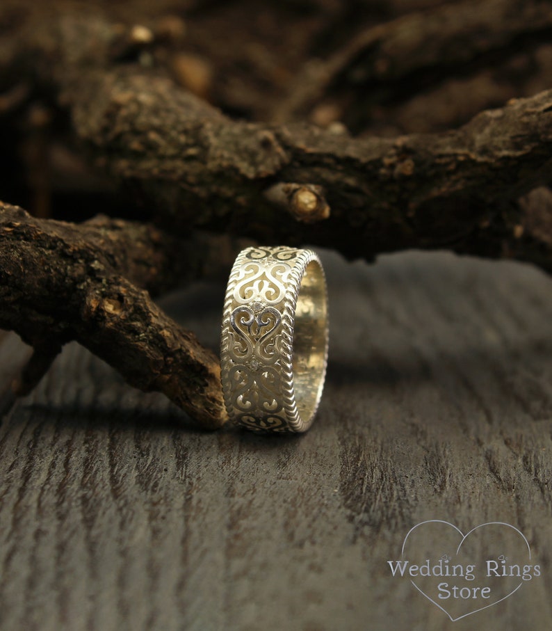 May include: A silver wedding band with a delicate, intricate design featuring a repeating heart pattern. The band is shown on a wooden surface.